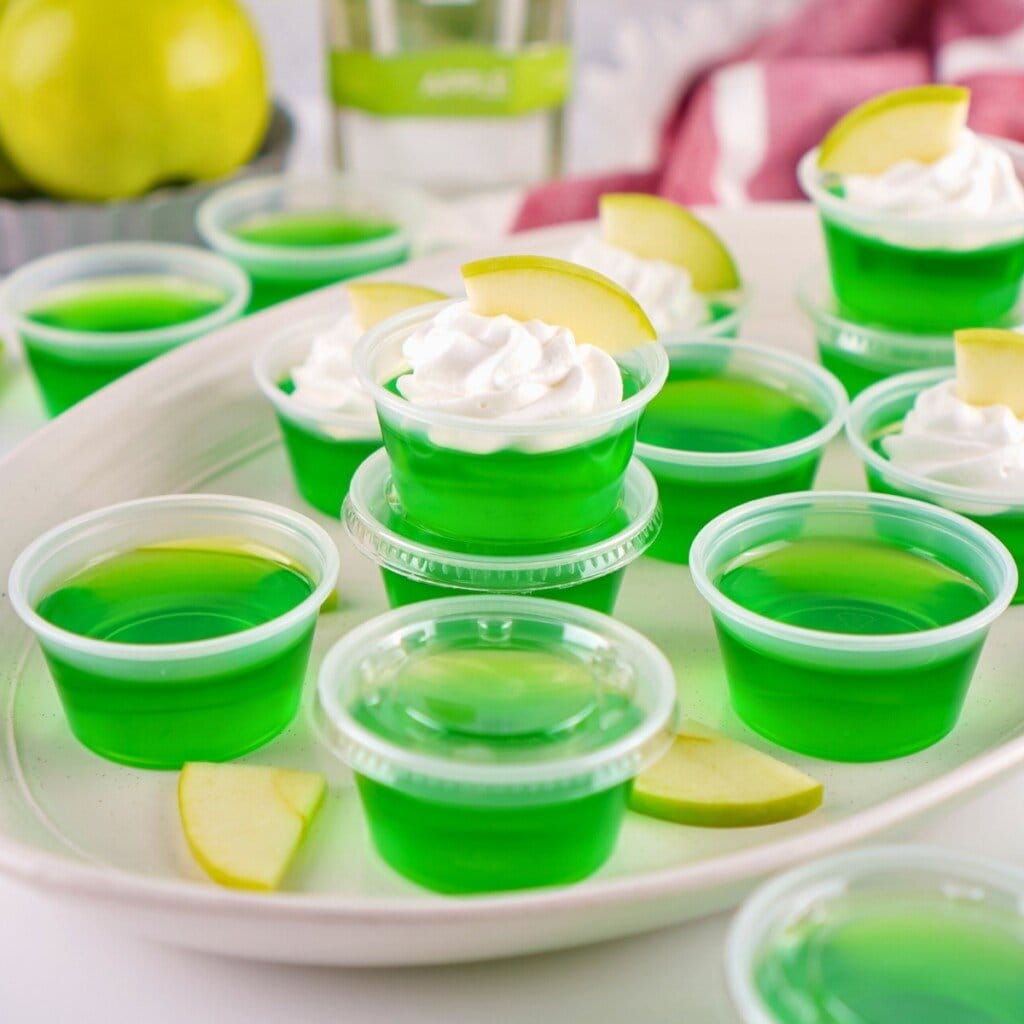 Green apple Jello shots in small cups, some topped with whipped cream and apple slices.