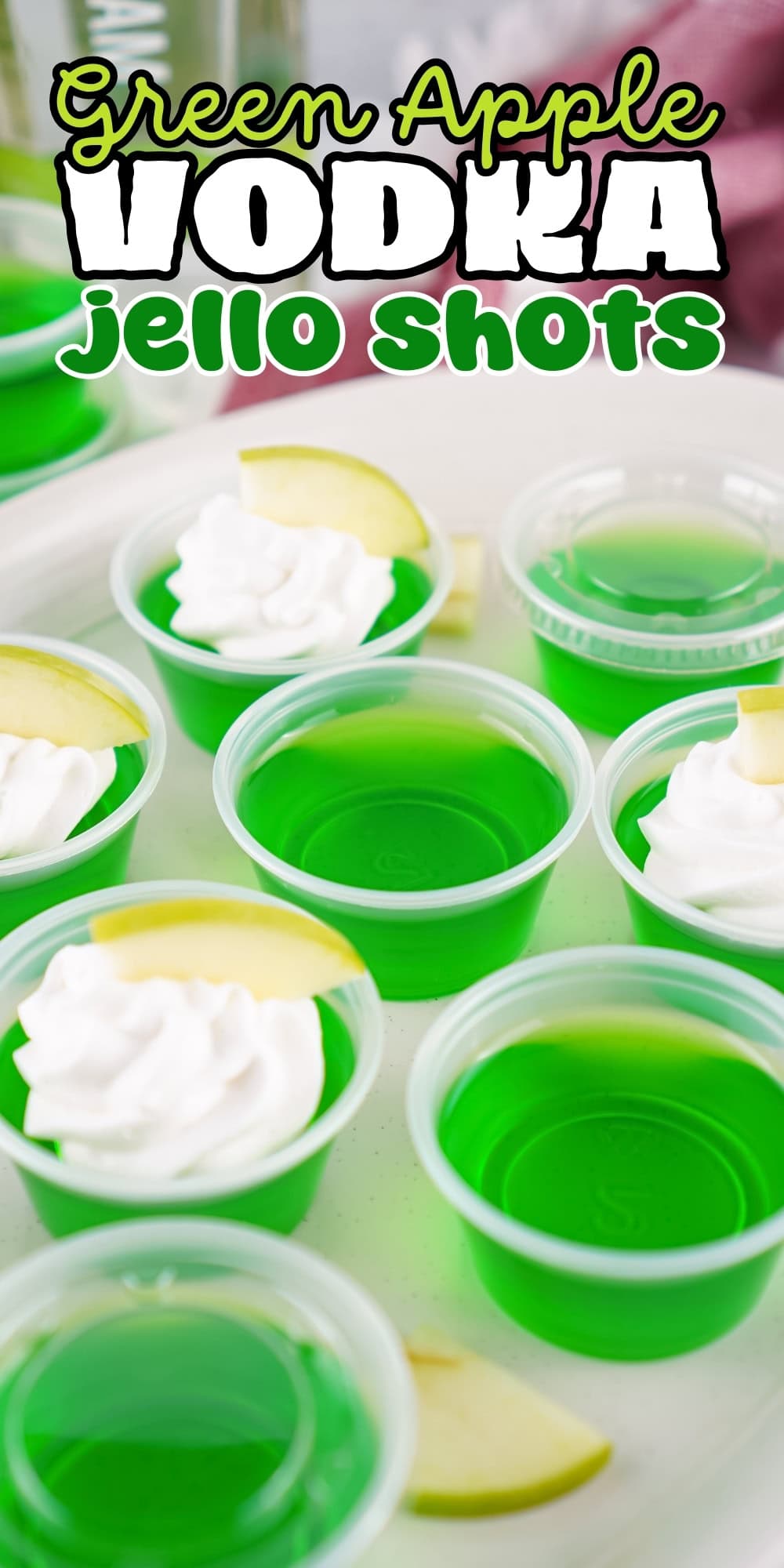 Green apple vodka jello shots in cups, some topped with whipped cream and apple slices.