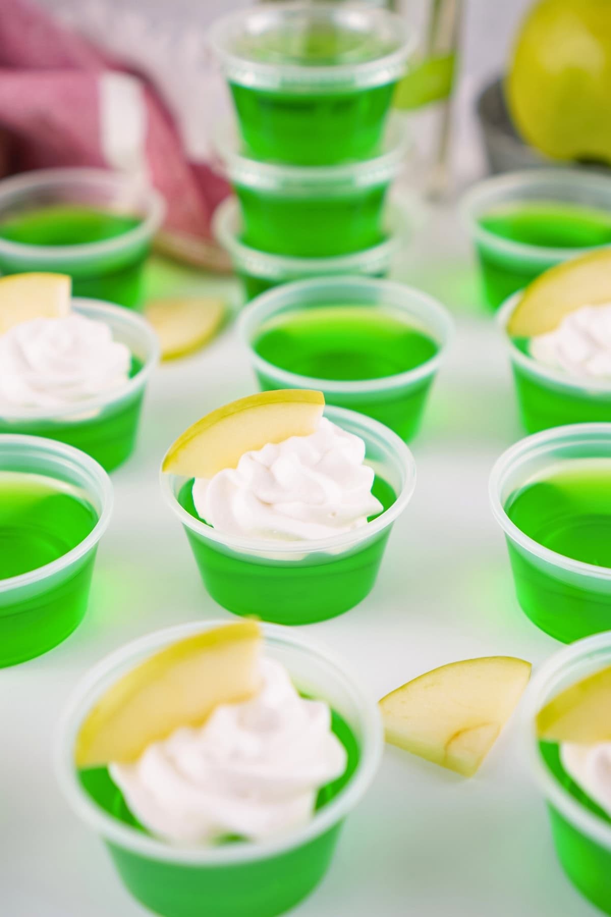 Green gelatin shots in plastic cups, some topped with whipped cream and apple slices.