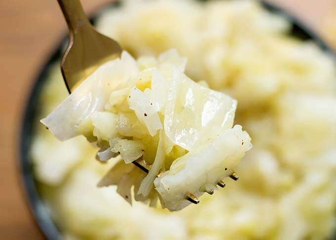 Fork holding a bite of cooked cabbage with pepper, over a bowl of cabbage.