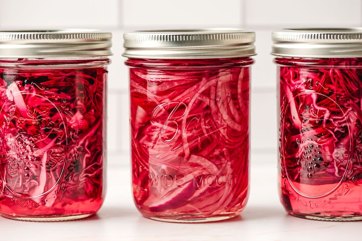 Three glass jars filled with pickled red onions on a white countertop.