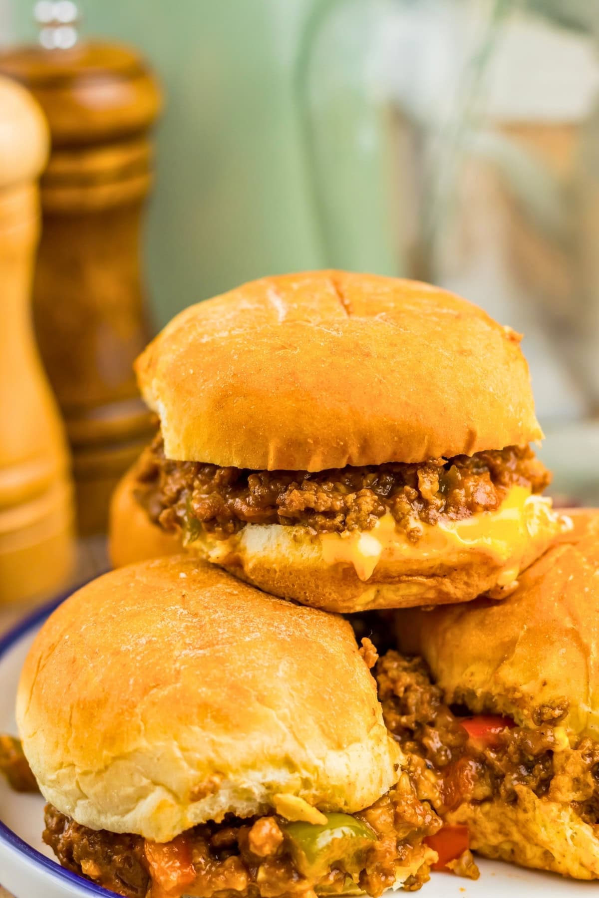 Three sloppy joe sandwiches stacked on a plate with blurred condiments behind them.