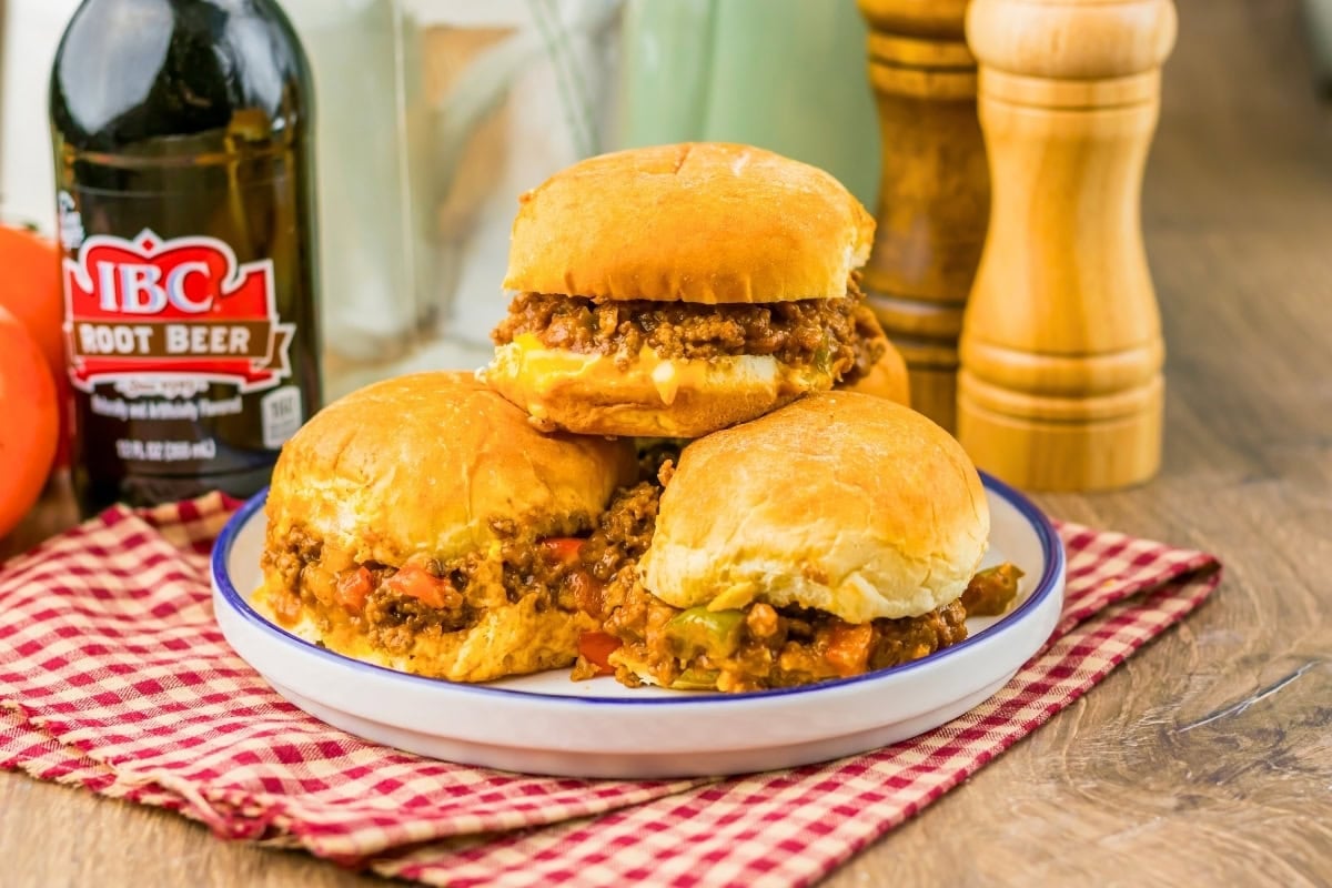 Three sloppy joe sandwiches on a plate next to a root beer bottle and condiments.