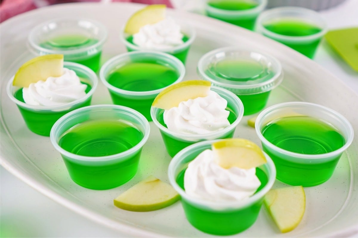 Green gelatin shots in cups, some topped with whipped cream and apple slices.