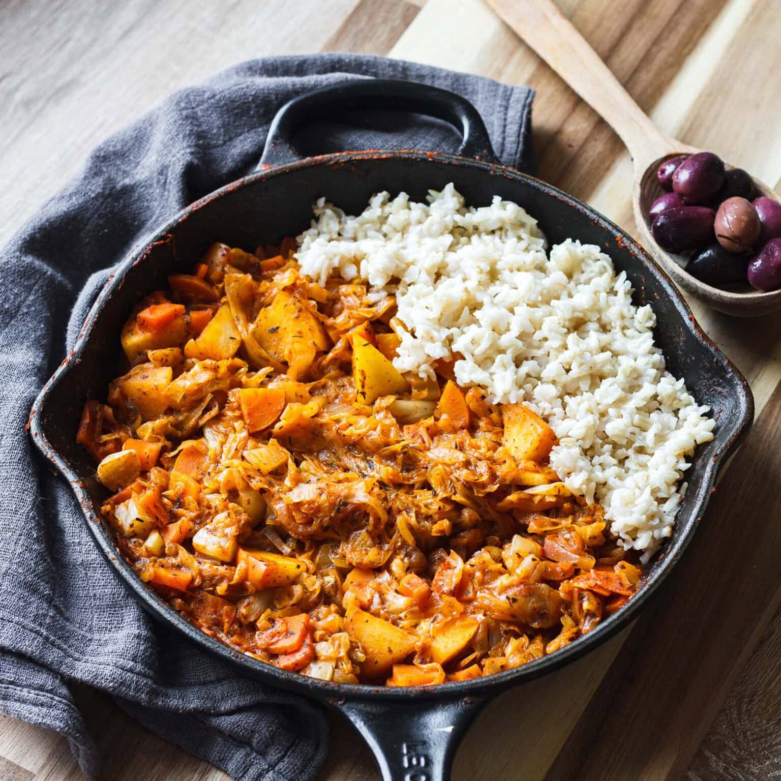A skillet with rice and a cooked vegetable stew, next to a wooden spoon with olives.