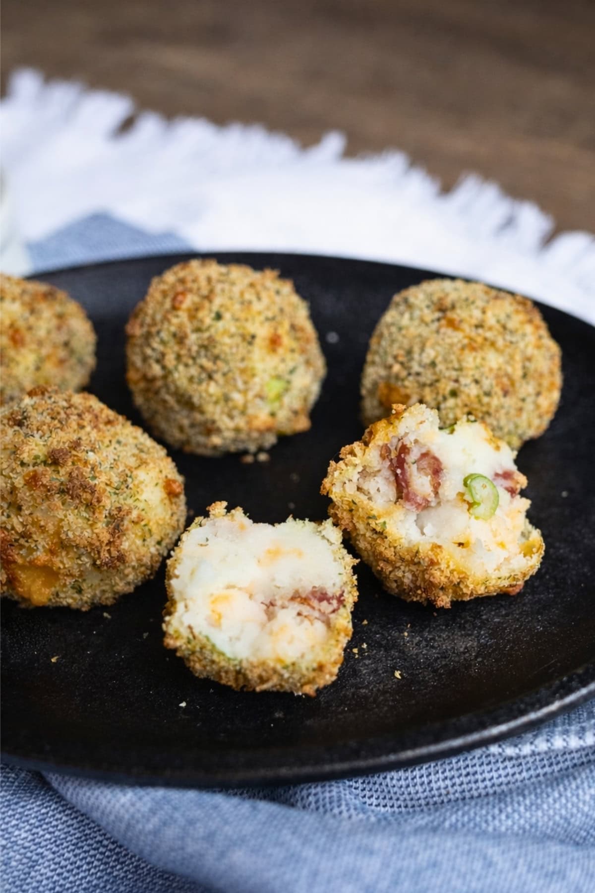 Five breaded mashed potato balls on a black plate, one cut open to show filling.