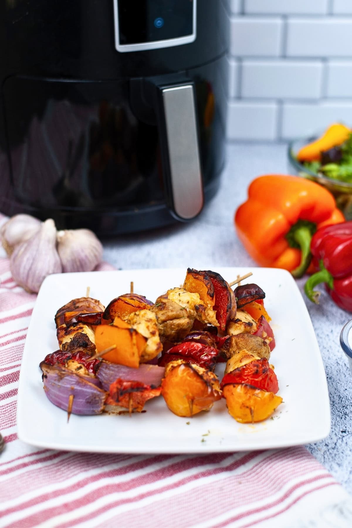 Chicken and vegetable skewers on a white plate in front of an air fryer.