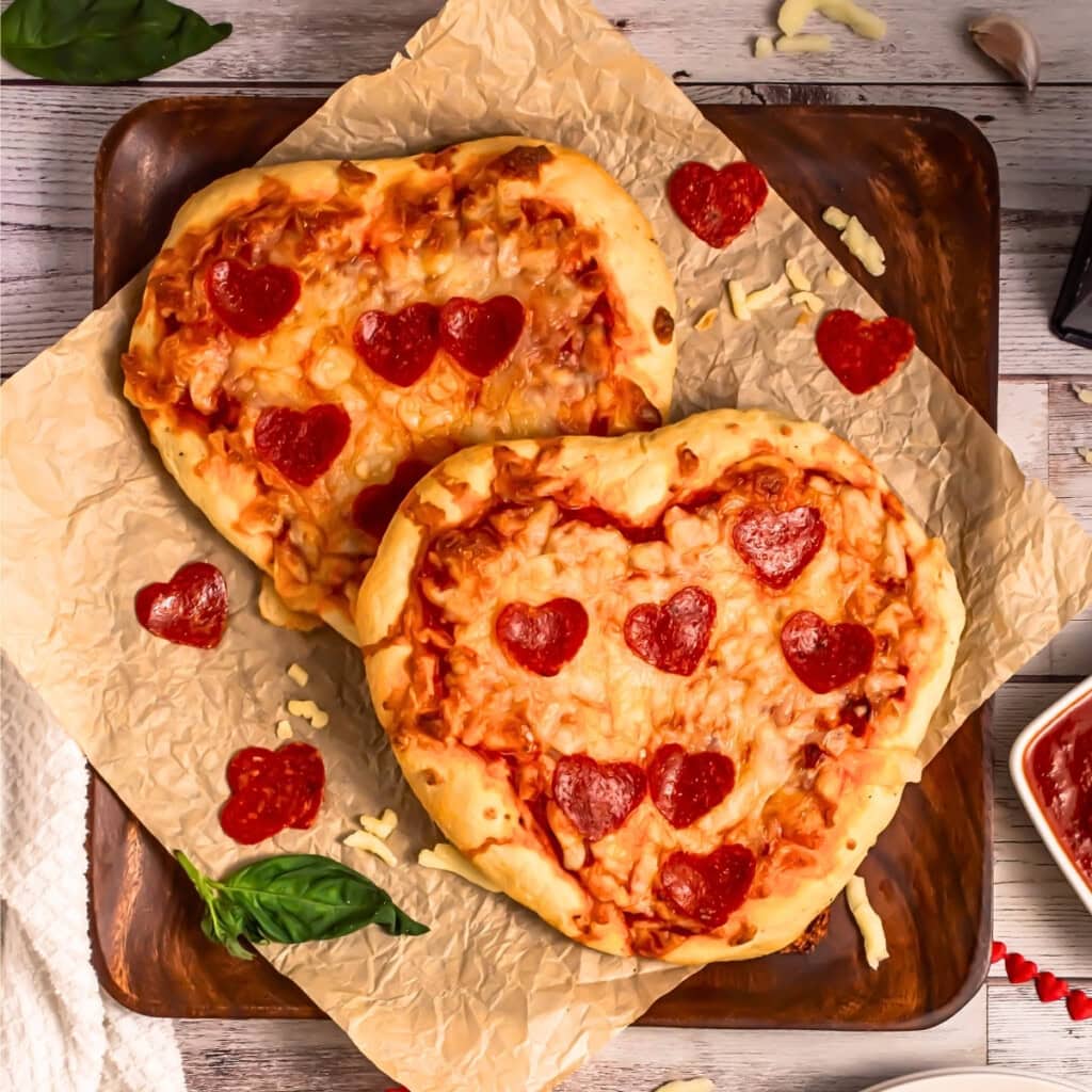 Two heart-shaped pizzas topped with heart-shaped pepperoni on parchment paper.