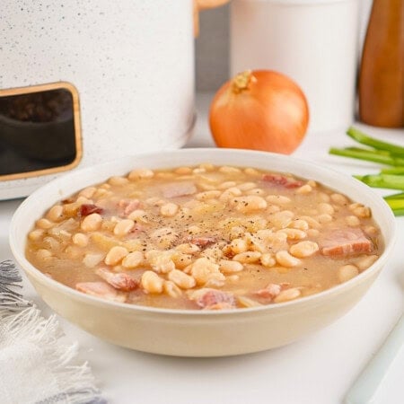 Bowl of white bean soup with ham, black pepper, and an onion in the background.