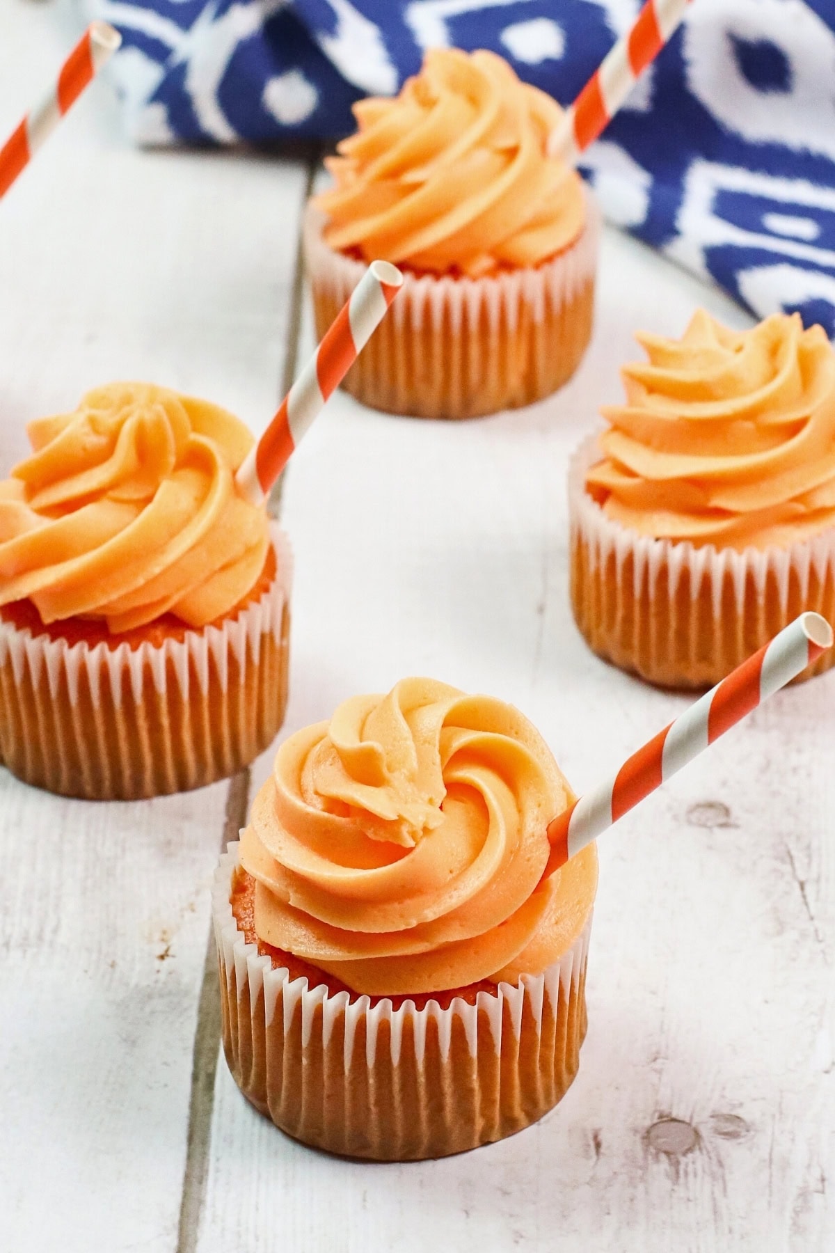 Four orange-frosted cupcakes with striped straws on a white wooden surface.