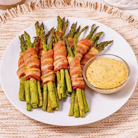 Bacon-wrapped asparagus bundles on a plate with a side of dipping sauce.