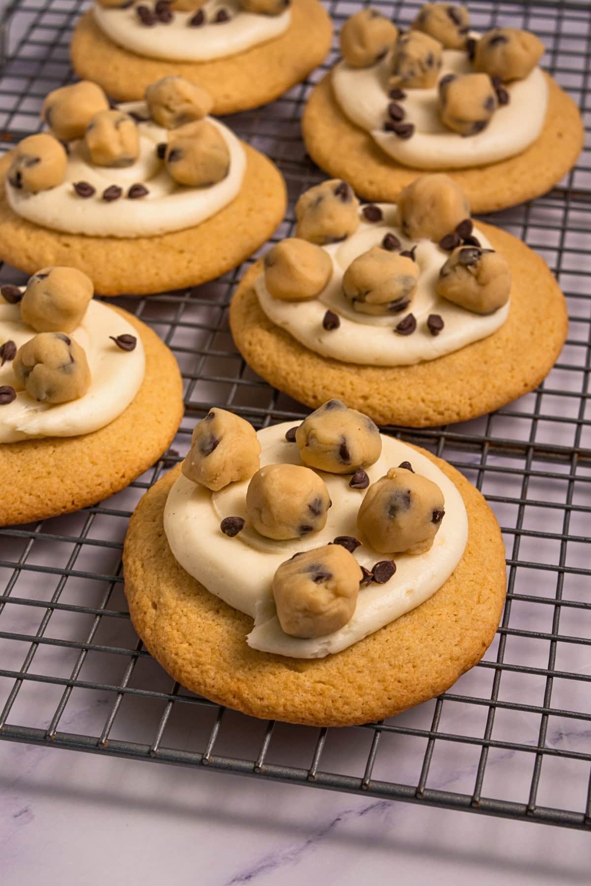 Cookies with frosting, topped with cookie dough balls and chocolate chips on a rack.