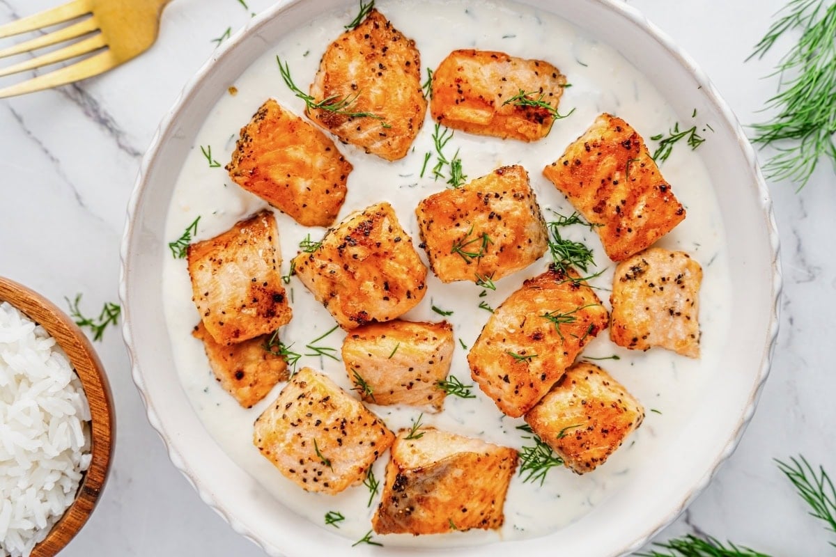Seared salmon bites in creamy sauce, garnished with dill, next to a bowl of rice.