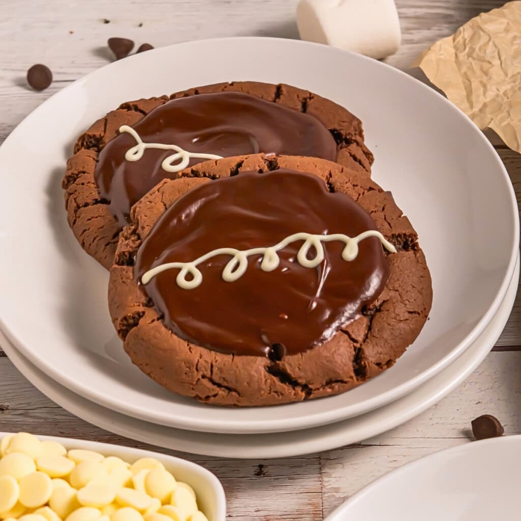Two chocolate cookies with glossy chocolate frosting and white icing squiggles on top.