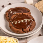 Two chocolate cookies with glossy chocolate frosting and white icing squiggles on top.