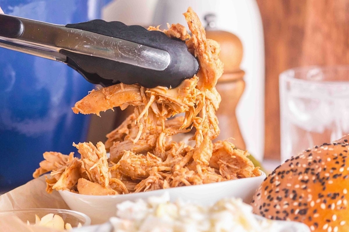 Tongs holding shredded barbecue chicken over a white bowl and sandwich bun.