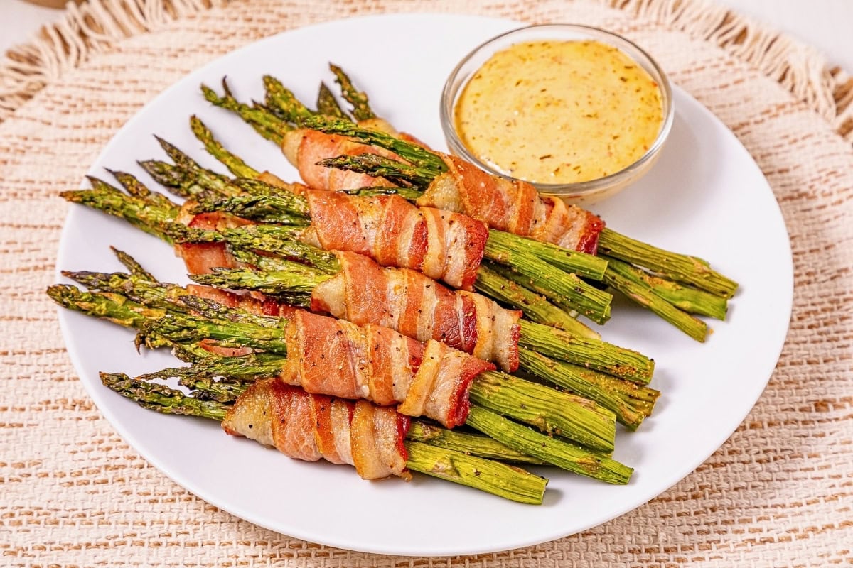 Bacon-wrapped asparagus bundles on a plate with a side of dipping sauce.