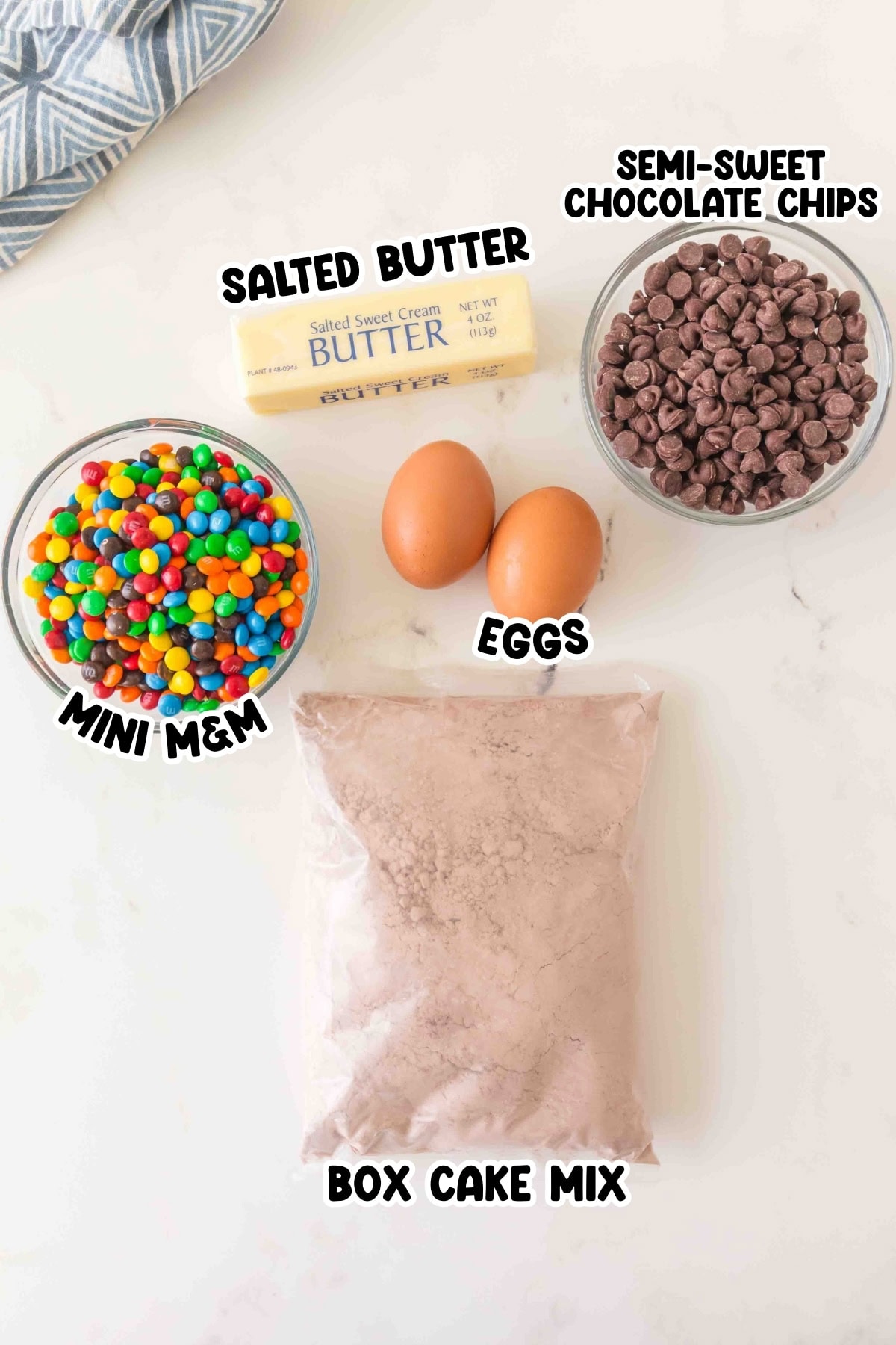 Box cake mix, mini MMs, two eggs, butter, and chocolate chips on a counter.