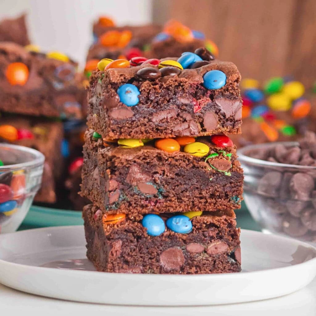 Three chocolate brownies with colorful candy pieces stacked on a white plate.