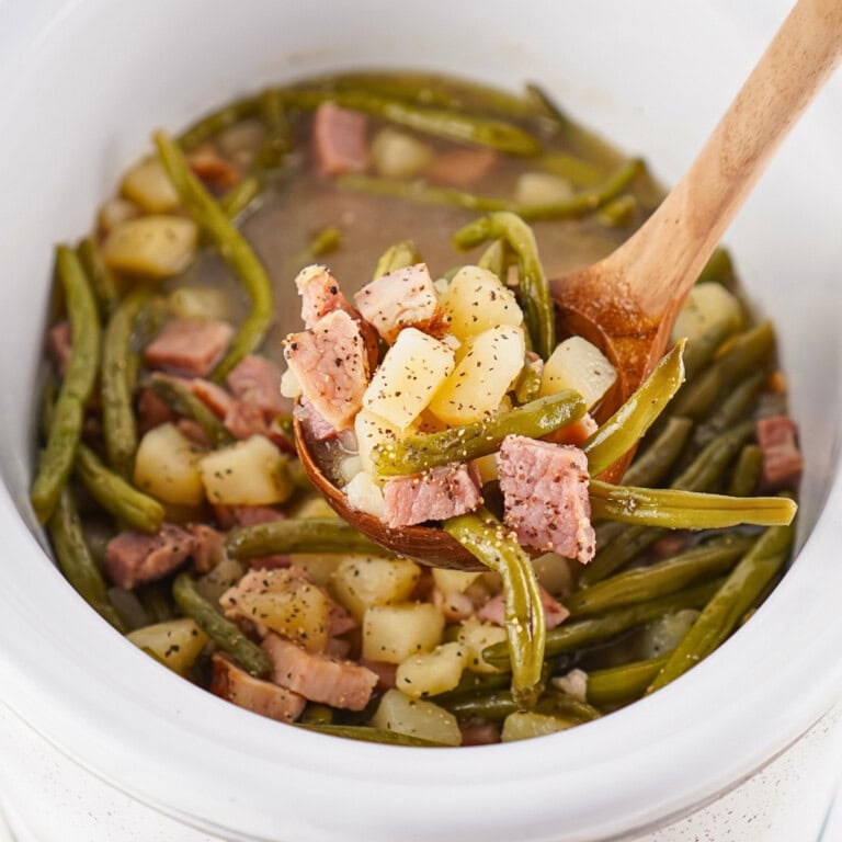 Wooden spoon lifting ham, green beans, and potatoes from a slow cooker.