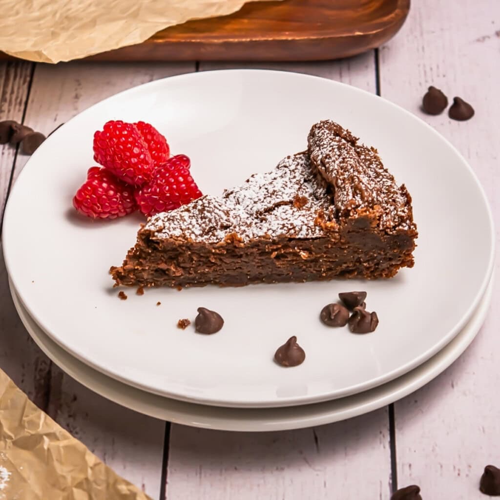 A slice of chocolate cake with raspberries and chocolate chips on a white plate.
