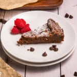 A slice of chocolate cake with raspberries and chocolate chips on a white plate.