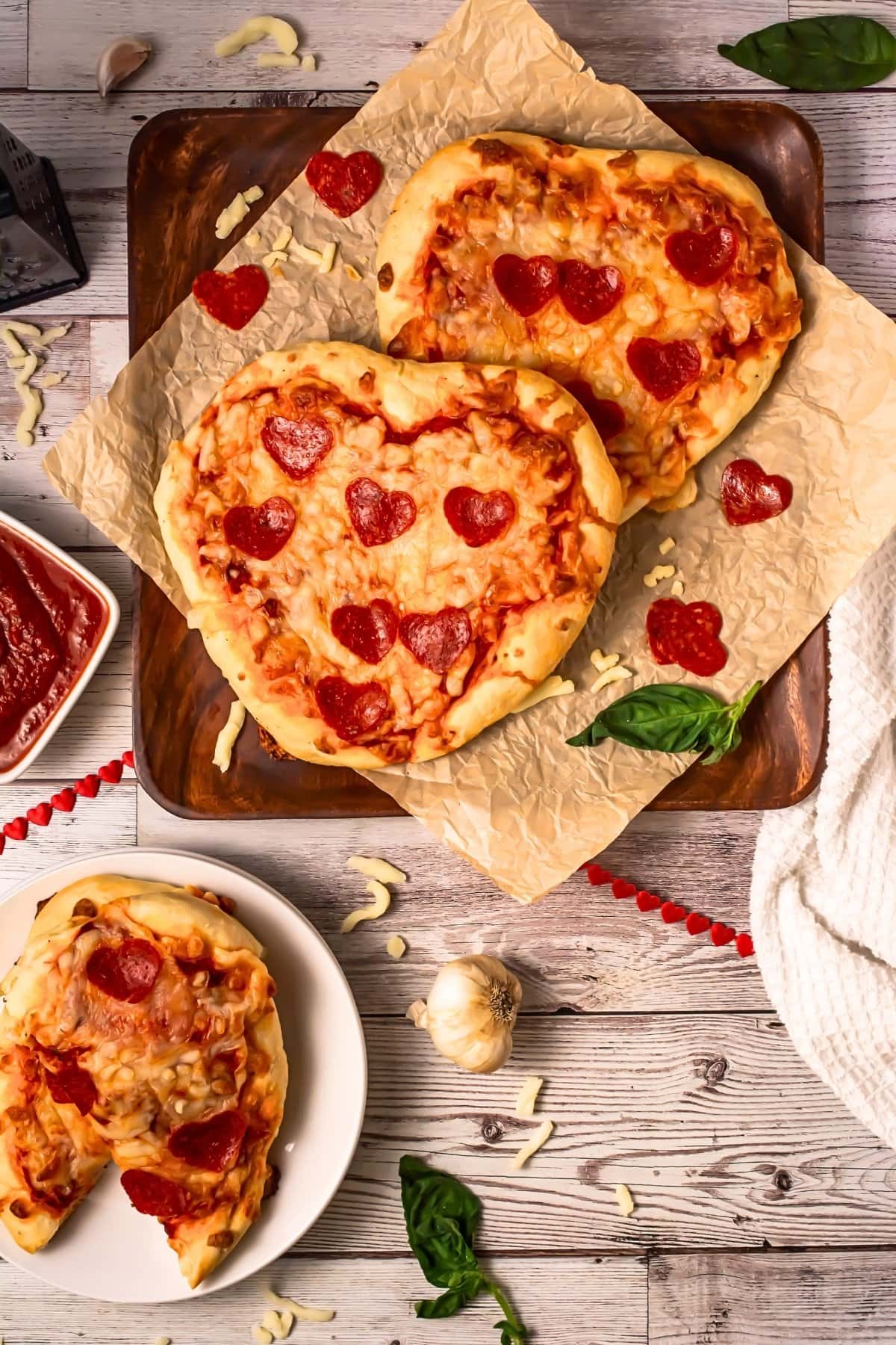 Heart-shaped pepperoni pizzas on parchment paper, with basil and sauce nearby.