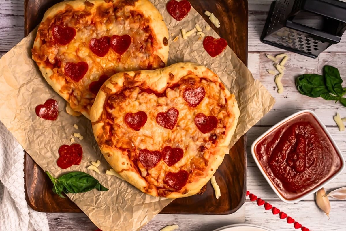 Two heart-shaped pizzas topped with heart-shaped pepperoni on parchment paper.