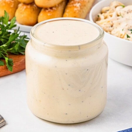 A jar of creamy Alfredo sauce with herbs, surrounded by breadsticks and parsley.