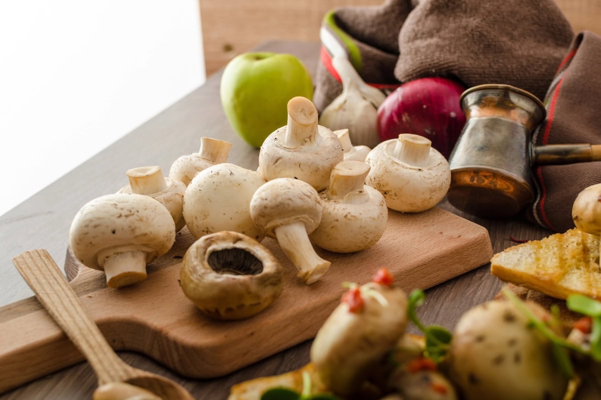 Fresh white mushrooms on a wooden board surrounded by various kitchen ingredients.
