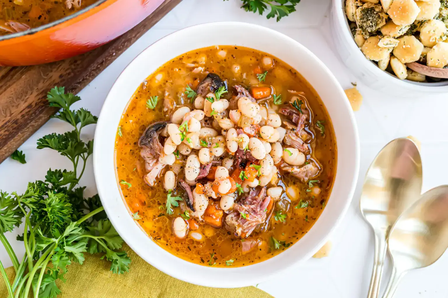 A bowl of bean soup with meat, carrots, herbs, and crackers on the side.