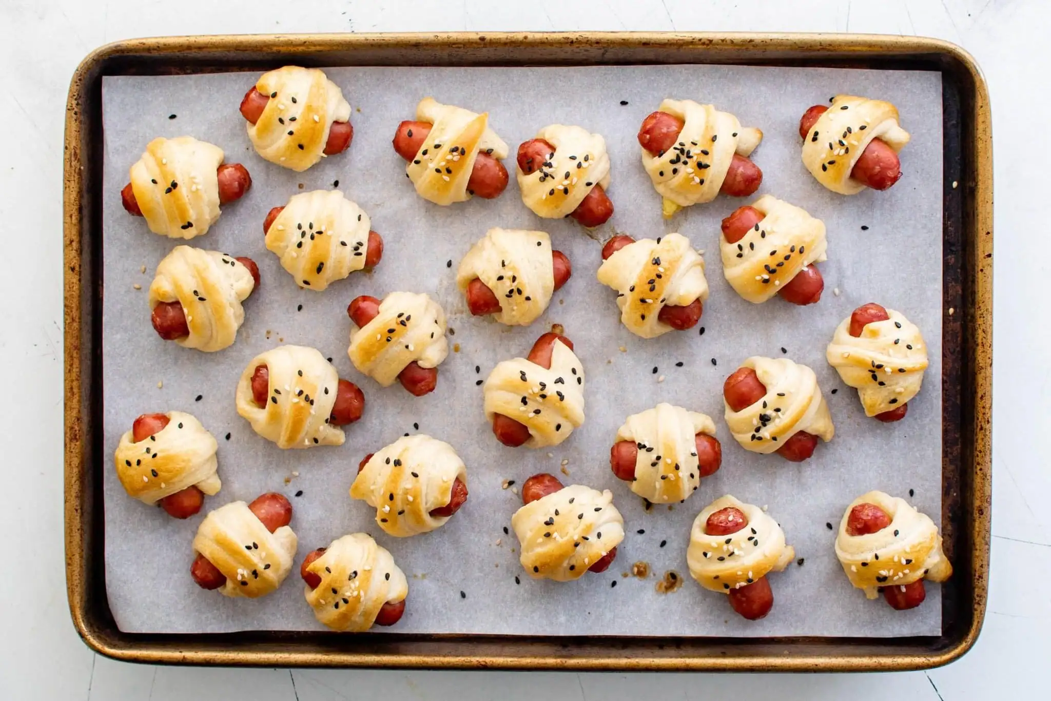 Mini pigs in a blanket with sesame seeds on a baking tray lined with parchment.