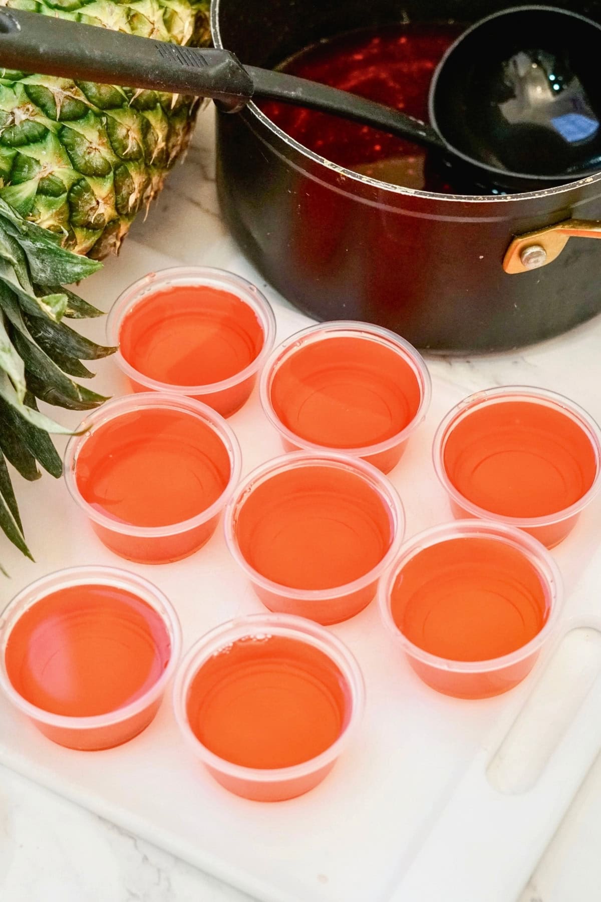 Nine small plastic cups filled with red liquid beside a pot and a pineapple.