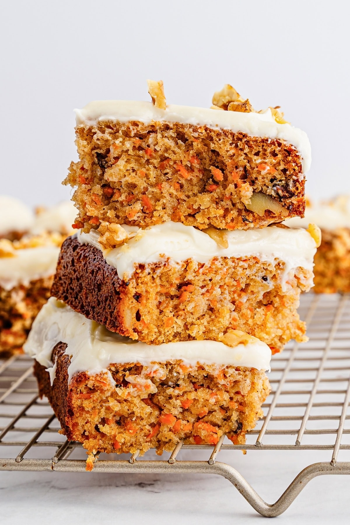 Three carrot cake bars with cream cheese frosting stacked on a cooling rack.