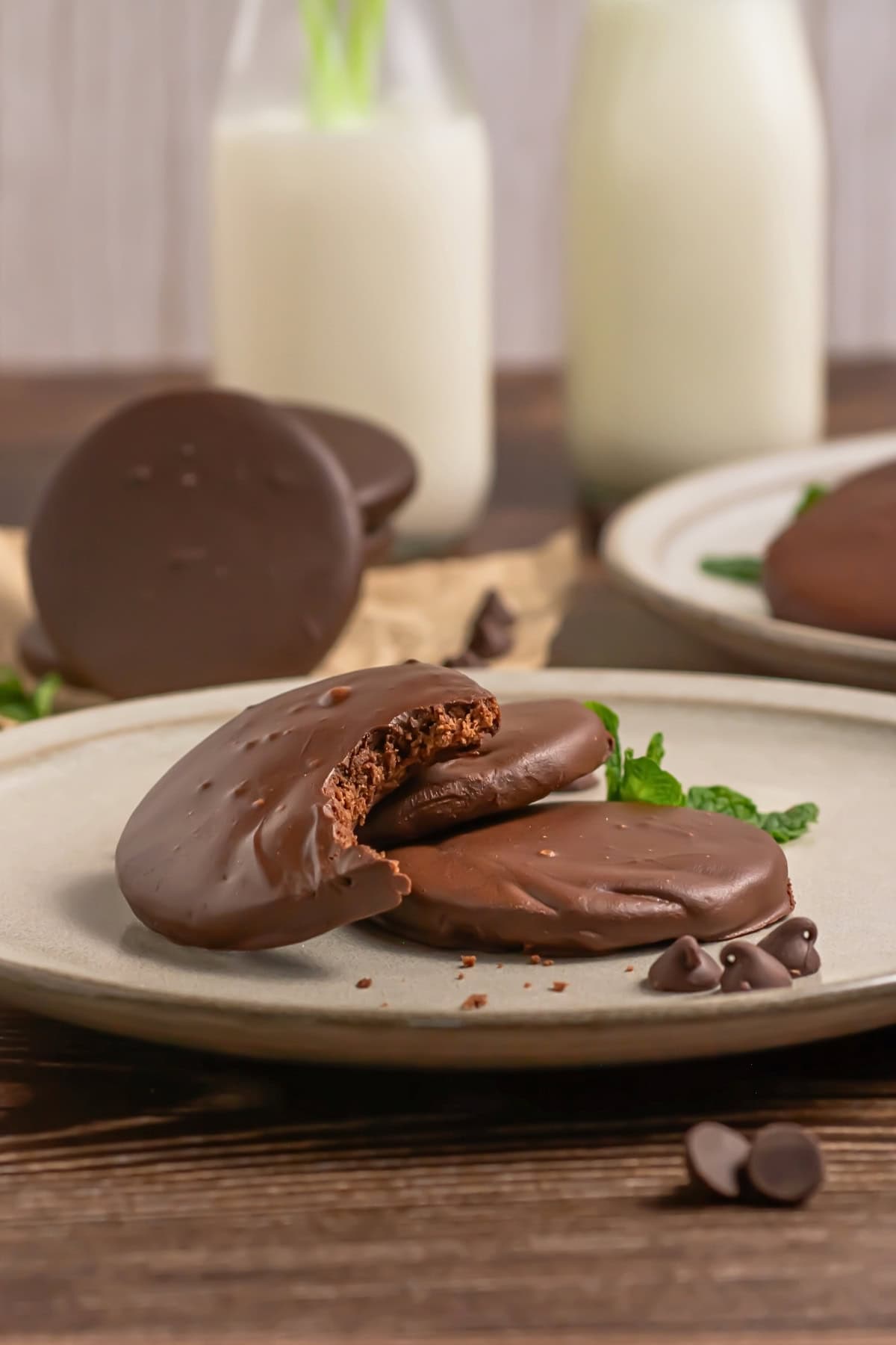 Chocolate-covered cookies on a plate, one with a bite taken, with milk in background.