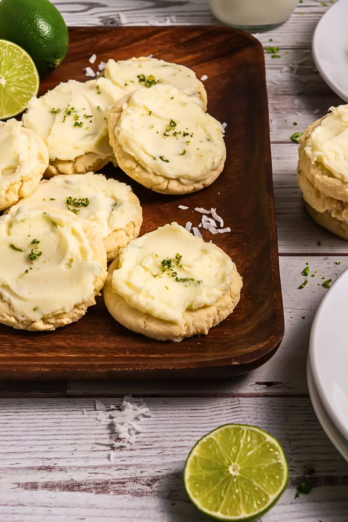Frosted cookies with lime zest on a wooden tray, surrounded by lime halves.