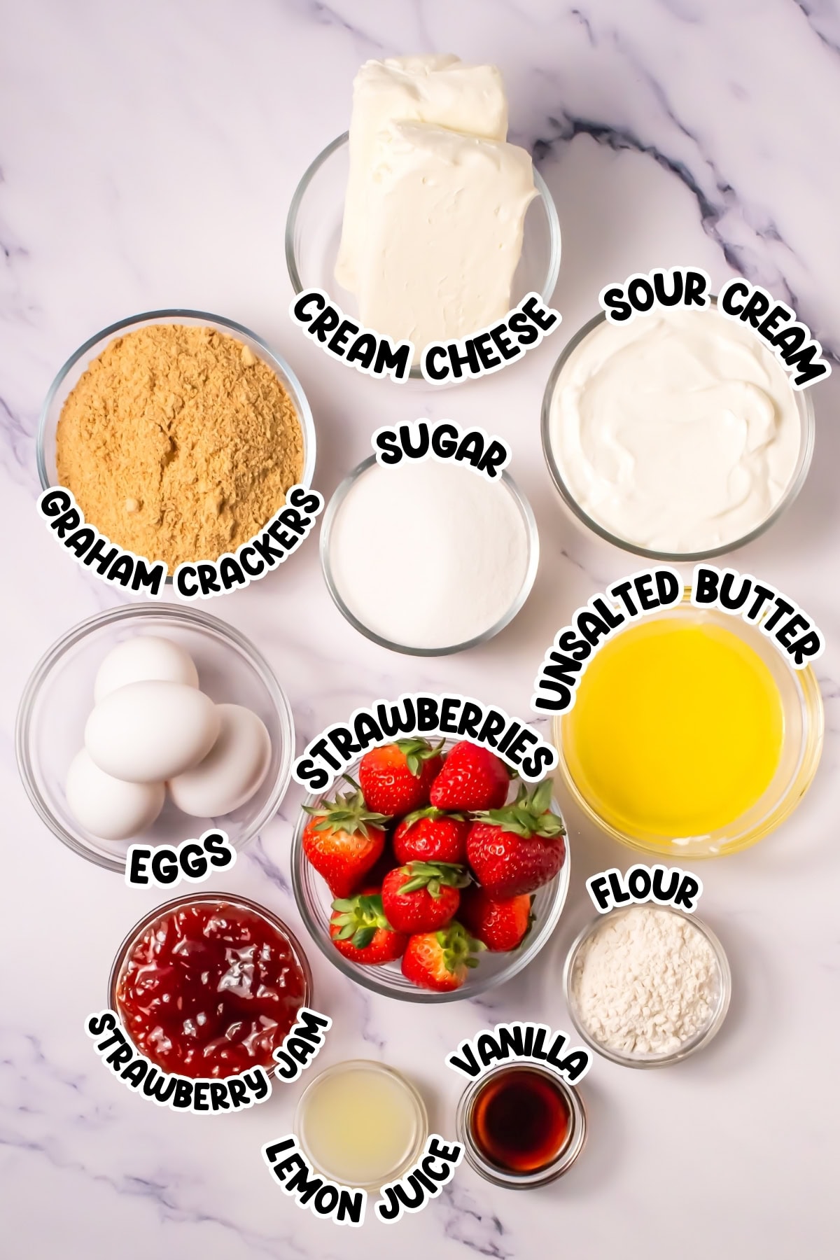 Ingredients for strawberry cheesecake, labeled in bowls on a white marble surface.