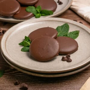 Three chocolate-covered cookies with mint leaves on a plate, with more cookies behind.