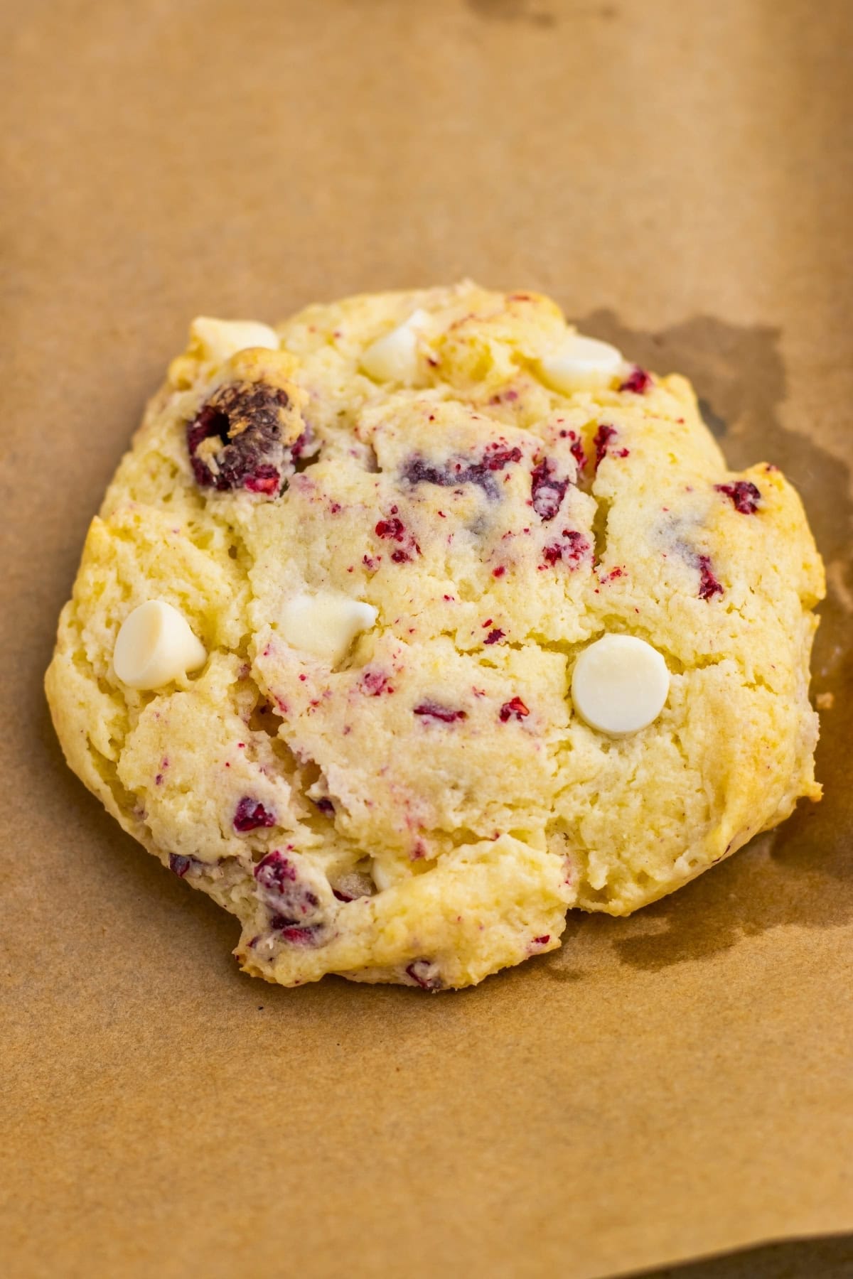 A cookie with white chocolate chips and cranberries on brown parchment paper.