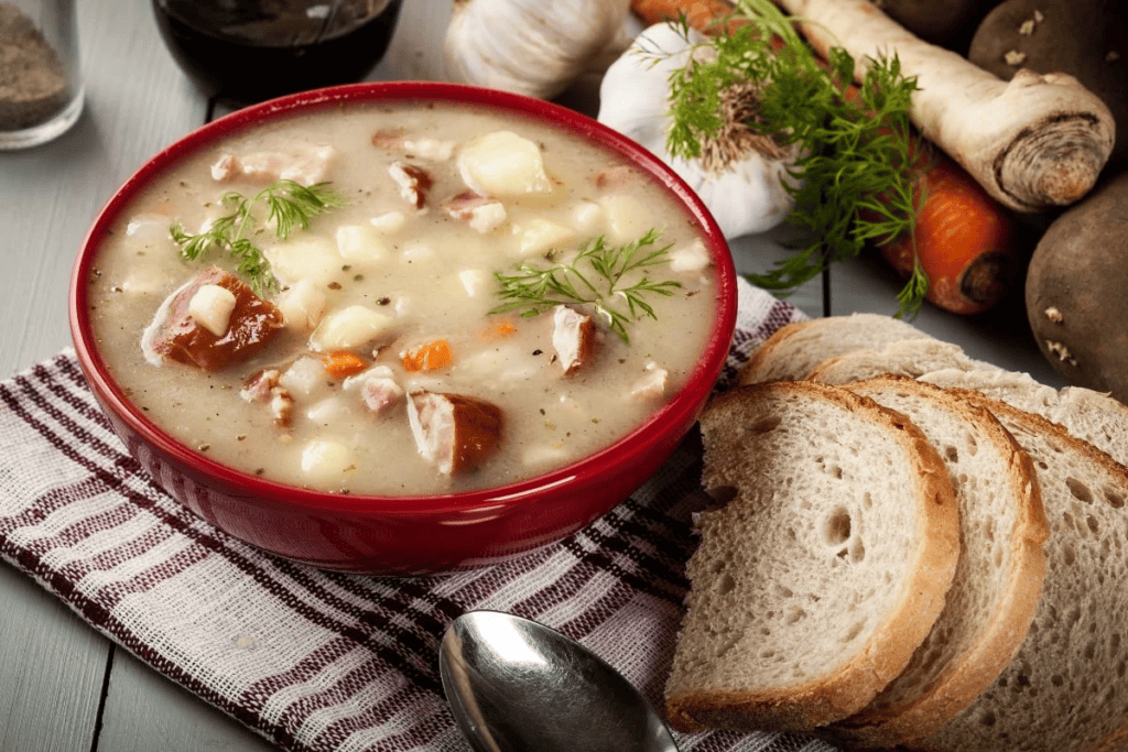 A bowl of soup with meat and vegetables, next to sliced bread and fresh vegetables.