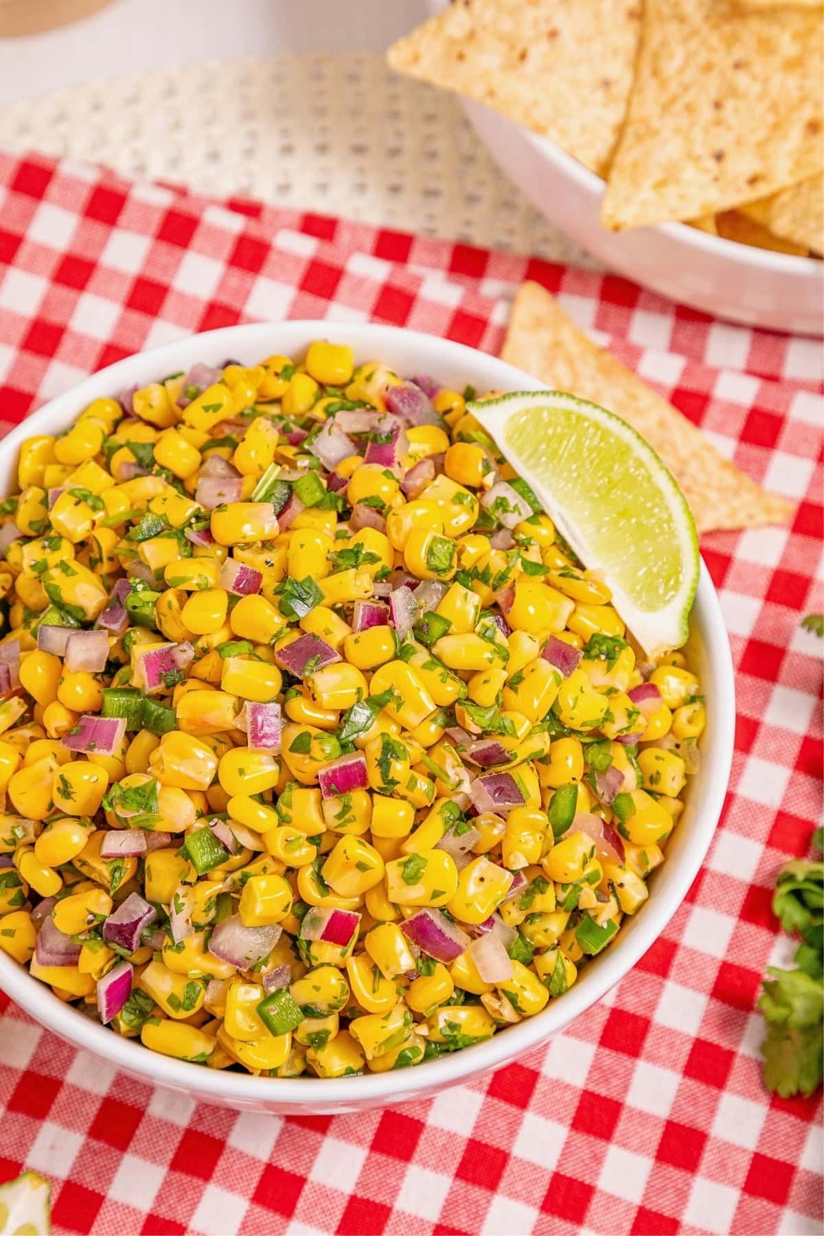 Bowl of corn salad with red onion, herbs, and a lime wedge on a red checkered cloth.