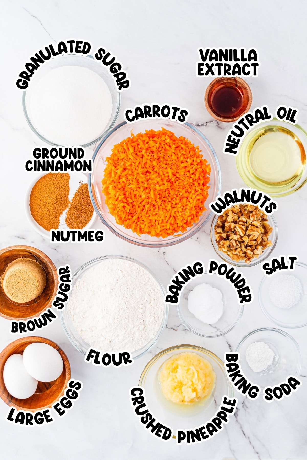 Top view of labeled carrot cake ingredients in bowls on a white surface.