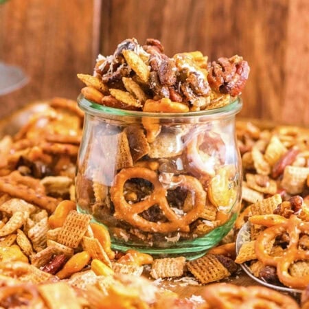A glass jar filled with snack mix including pretzels, nuts, and cereal pieces.