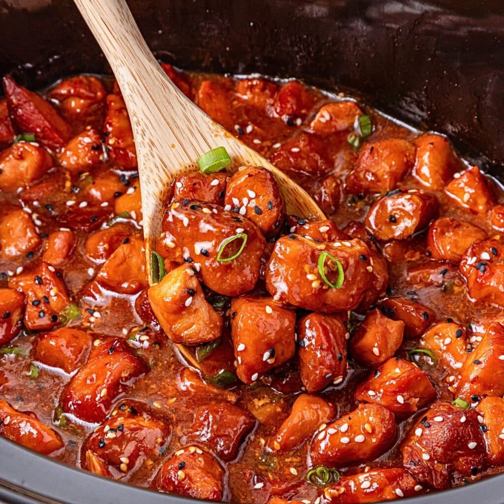 Wooden spoon scooping glazed chicken bites with sesame seeds and green onions.