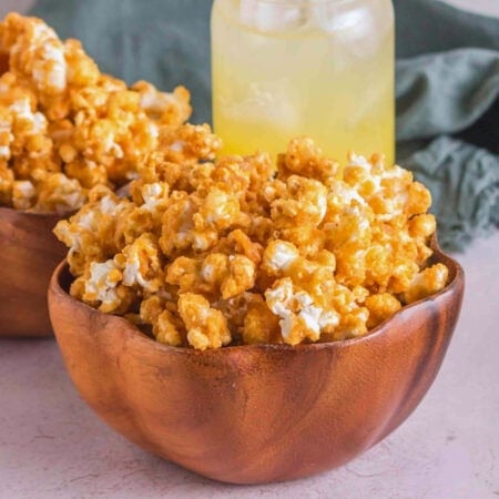 Caramel popcorn in a wooden bowl with a drink in the background.