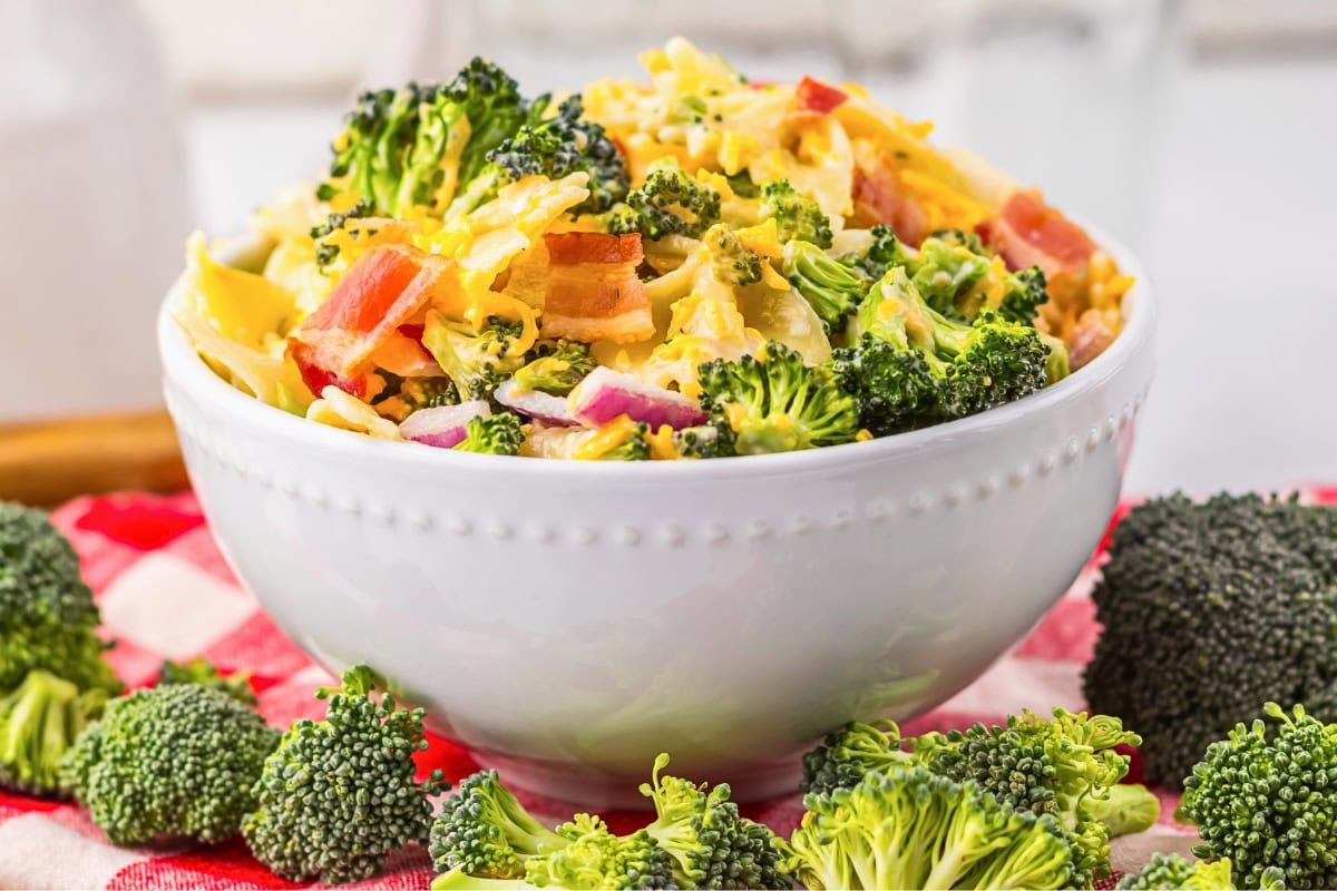 A bowl of broccoli salad with bacon, cheese, and red onions on a tablecloth.