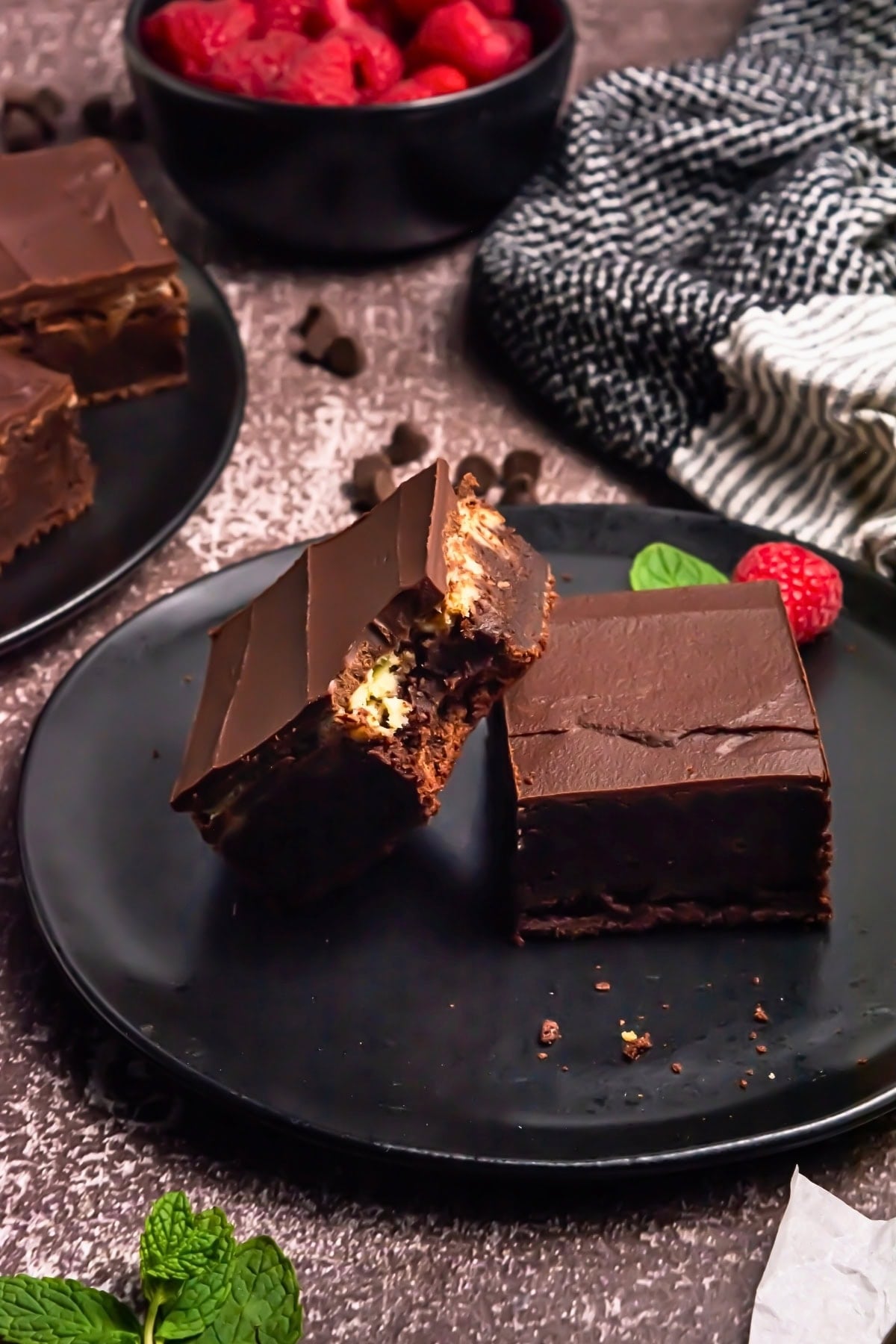 Chocolate brownies on a black plate, with mint leaves and raspberries nearby.