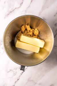 Metal mixing bowl with butter, sugar, and brown sugar on a marble surface.