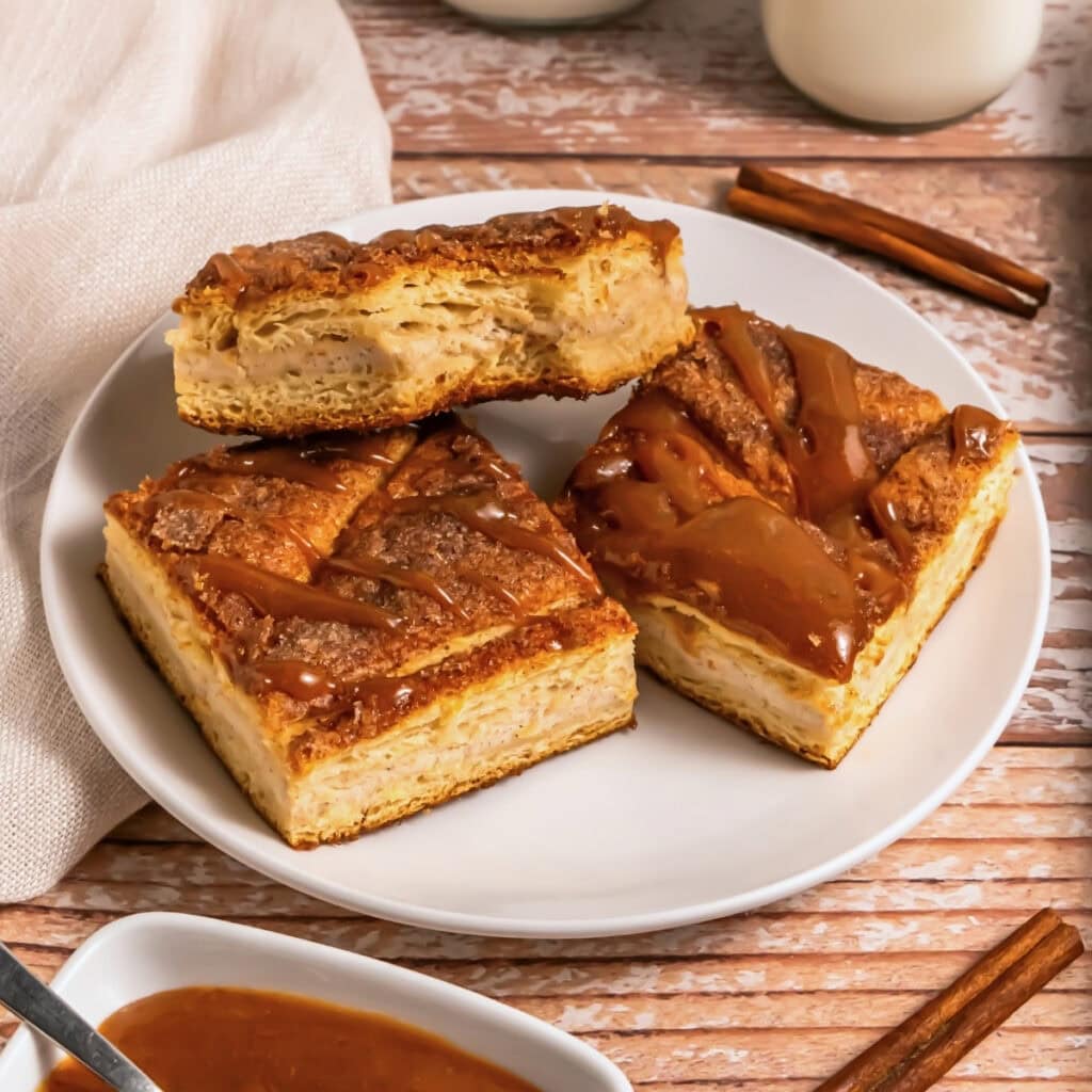 Three caramel-drizzled pastry squares on a white plate with cinnamon sticks.