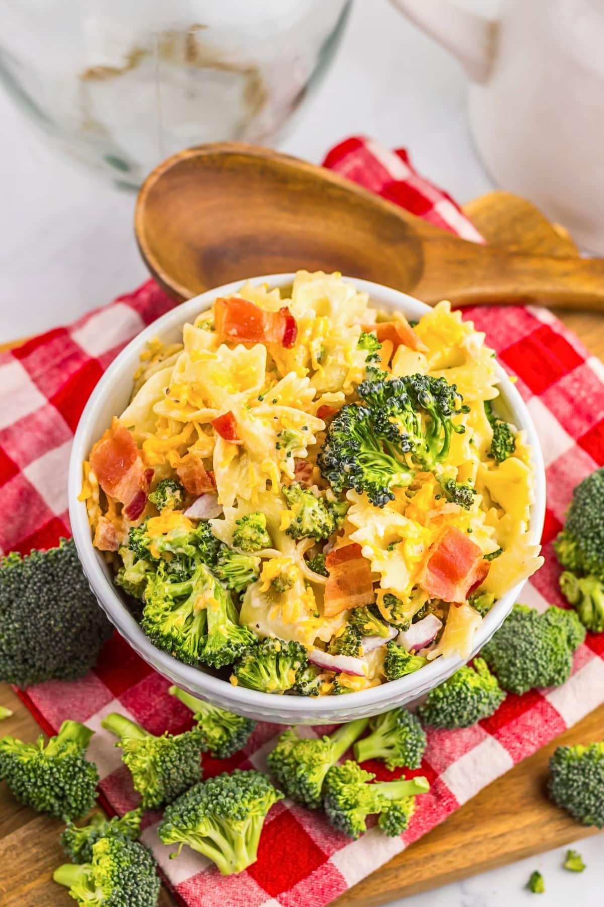 Bowl of pasta salad with broccoli and tomatoes on a red checkered cloth.